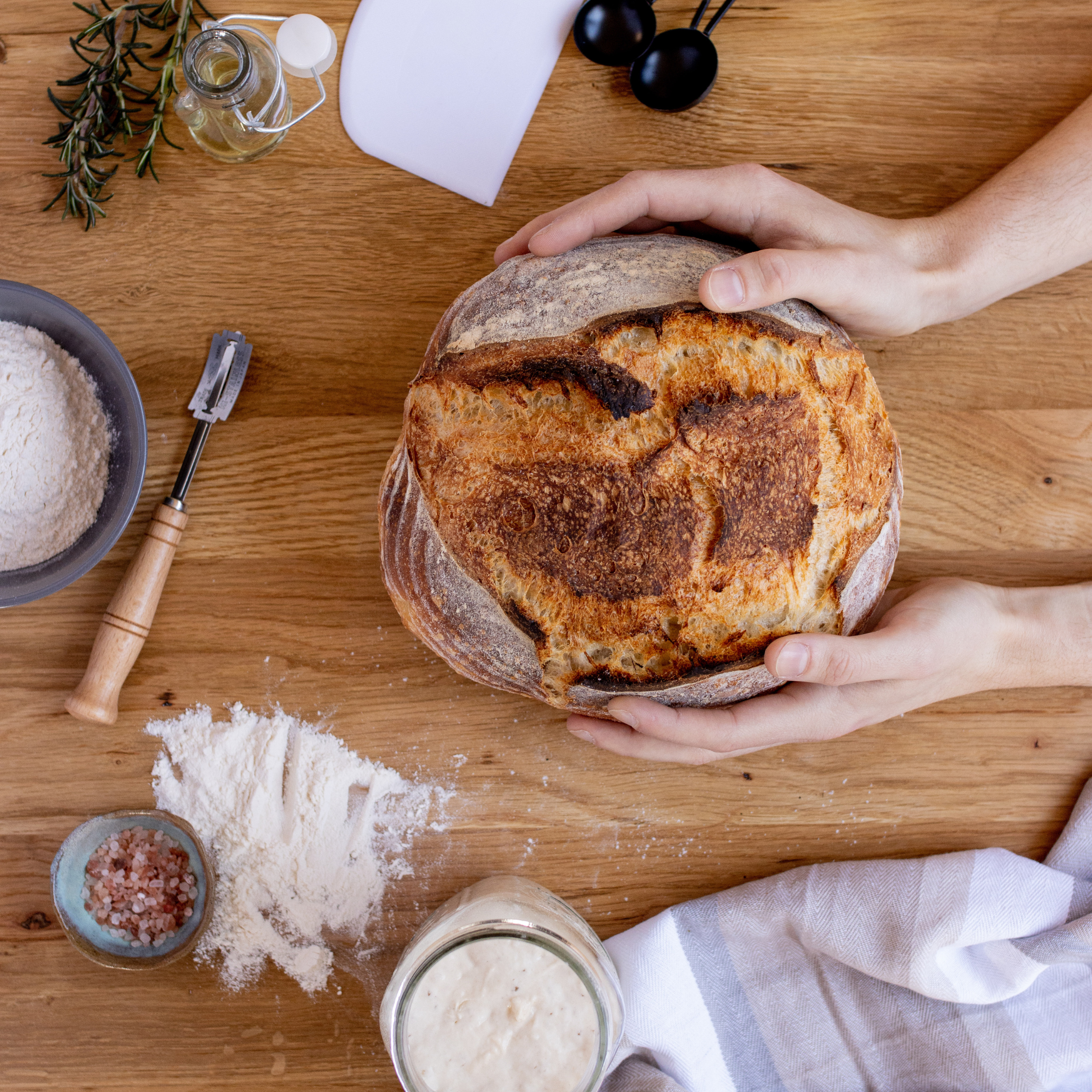 Fresh sourdough loaf on wooden bench with bread lame, flour, salt, dough scraper and measuring utensils
