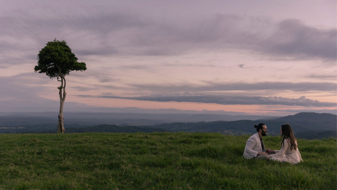 Dan and Candice Kennedy, Co-creators of Eden Tree Eco sitting together hand in hand in grass with a back drop of One Tree Hill under a violet dusk sky.