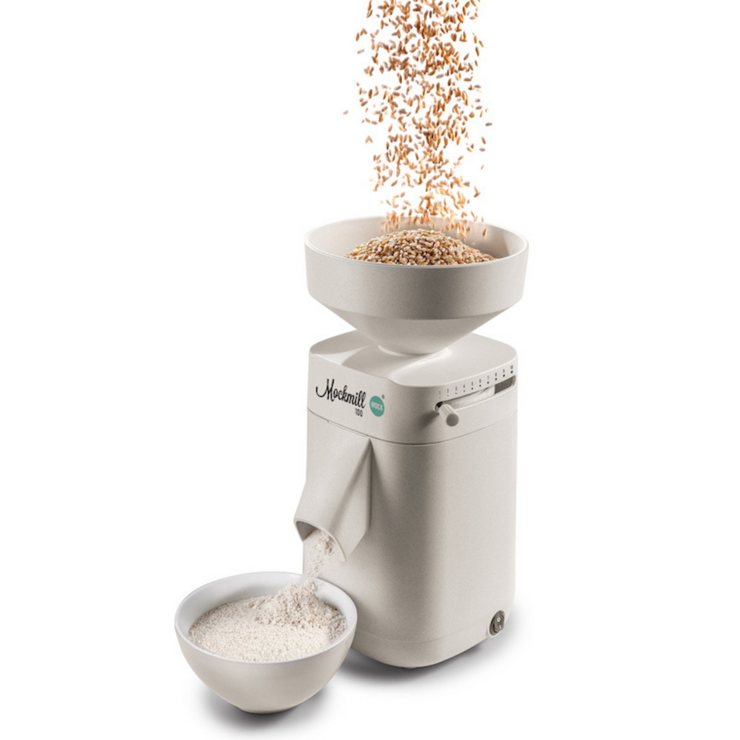 A Mockmill 100 MOCK turned on and operating on a white background with grain / wheat being poured into the top (hopper) and producing flour into a white bowl
