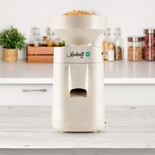 A Mockmill 100 MOCK grain and flour mill sitting on a white kitchen bench in an eco kitchen with indoor plants, food storage jars and condiments