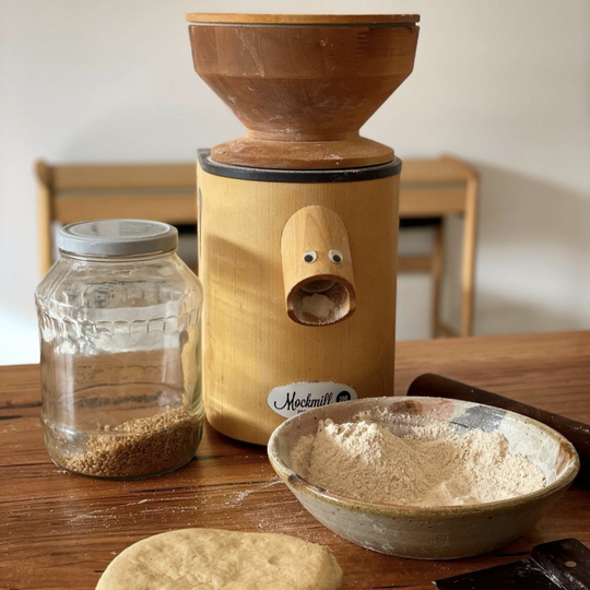 The Mockmill 200 PRO professional MOCK wooden flour and grain mill for personal use on a wooden kitchen table with a jar of brown rice and freshly milled ground brown rice flour and some dough